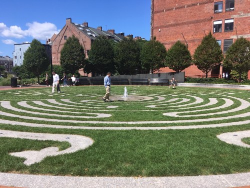 A circular stone labyrinth in a park, with people walking and a small fountain in the center, surrounded by greenery.
