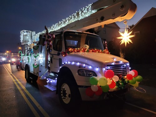 A decorated bucket truck adorned with colorful lights and festive ornaments, parked on a street at dusk.