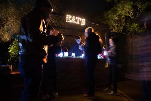 A nighttime scene at a food stand with people interacting and a sign that reads "EAT'S" illuminated above.