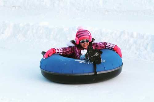 A child in winter attire enjoys tubing down a snowy slope, wearing sunglasses and a pink striped hat.