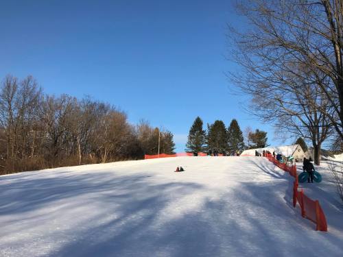 A snowy hill with people sledding, surrounded by trees and a clear blue sky. Orange fencing lines the slope.
