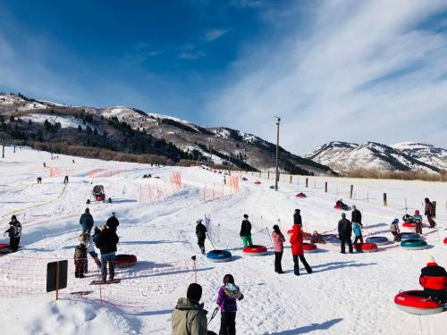 A snowy landscape with people tubing on a hill, surrounded by mountains and a clear blue sky.