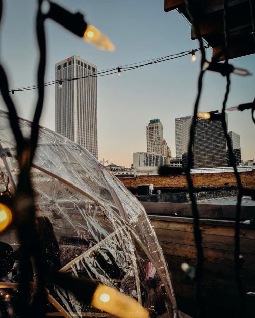 A clear dome with string lights in the foreground, overlooking city skyscrapers at sunset.