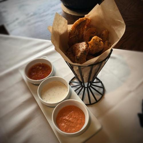 A wire basket filled with fried snacks, accompanied by three small bowls of dipping sauces on a white tablecloth.