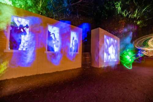 Colorful light projections of musicians on a wall, surrounded by vibrant swirls of light in a dark outdoor setting.