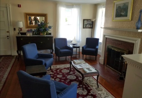 Cozy living room with blue chairs, a fireplace, a decorative mirror, and a patterned rug. Natural light from windows.