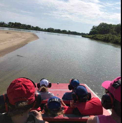 A group of people wearing headphones sits in a boat, facing a calm river with lush greenery on the banks.