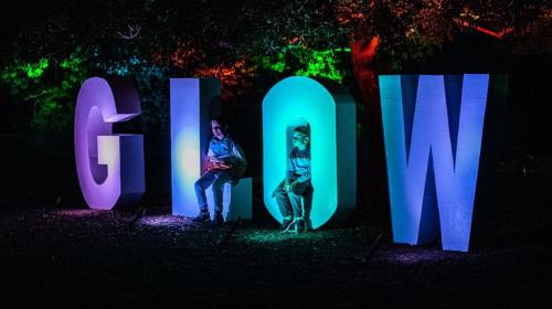 Two children sit beside large illuminated letters spelling "GLOW" in vibrant colors at night.