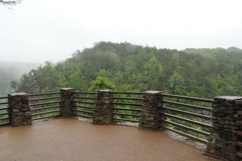 A misty view from a stone railing overlook, surrounded by lush green trees and foggy hills in the background.