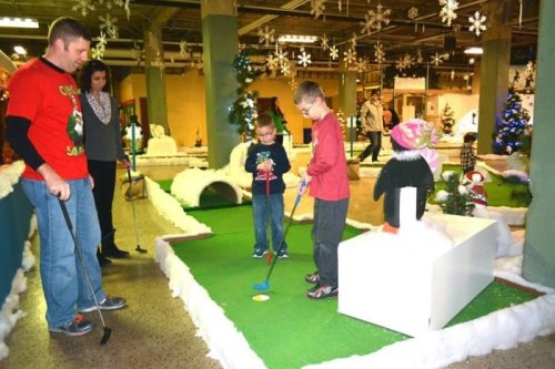 A family enjoys indoor mini-golf decorated for the holidays, with kids playing and adults watching in a festive setting.