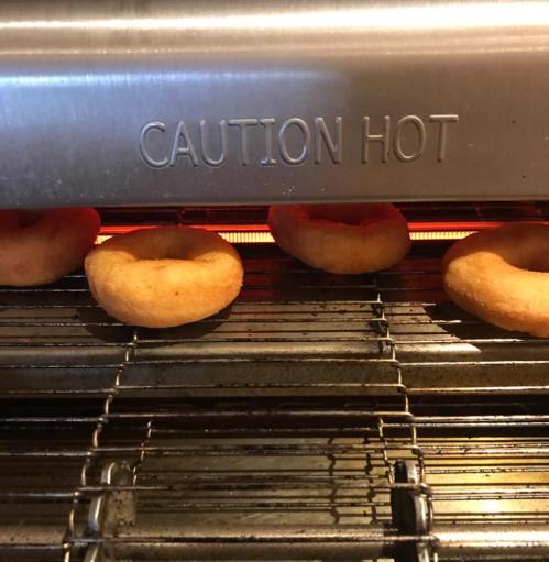 Freshly cooked donuts resting on a wire rack inside a hot oven, with a "Caution Hot" sign above.