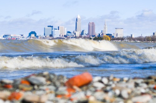 Waves crash on a rocky shore with a city skyline in the background under a clear blue sky.