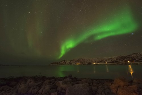 A stunning display of the Northern Lights over a calm lake, with rocky shores and distant hills illuminated by stars.