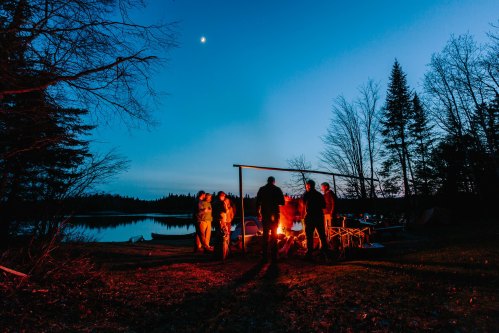 A group of people gathered around a campfire by a lake at dusk, with trees silhouetted against a blue sky.