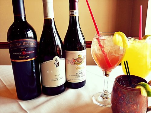 A selection of wine bottles and colorful cocktails on a table, featuring Merlot, Pinot Noir, and Barolo.