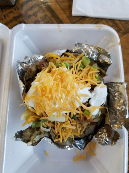 A loaded baked potato topped with shredded cheese, sour cream, and green onions, served in a takeout container.