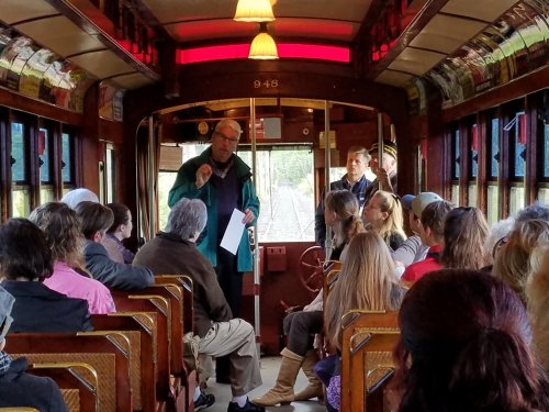 A group of people seated on a vintage train, listening to a speaker at the front of the car.