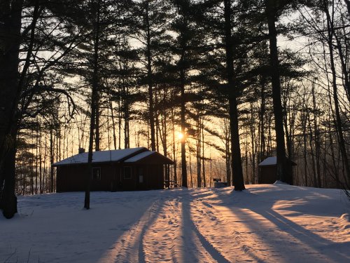 A serene winter scene with two cabins surrounded by snow-covered trees, illuminated by a warm sunset.