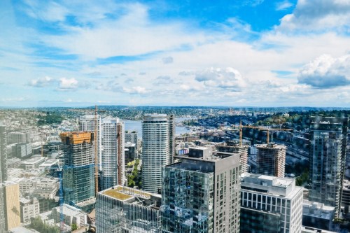 A panoramic view of a city skyline with modern buildings, water, and a blue sky dotted with clouds.