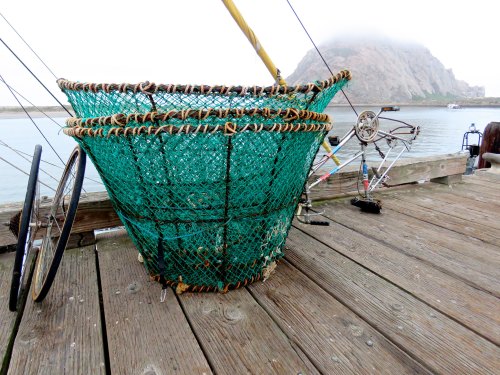 A green fishing basket on a wooden dock, with a foggy mountain and boats in the background.