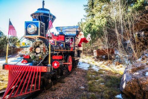 A festive train decorated for the holidays, with Santa waving from the front, surrounded by snow and greenery.