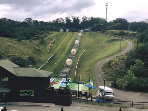 A grassy hill with a slide and inflatable balls, surrounded by trees and a building labeled "Outdoor Gravity Park."