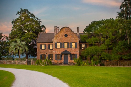 Historic brick house with large windows, surrounded by greenery and a gravel path, under a colorful sunset sky.