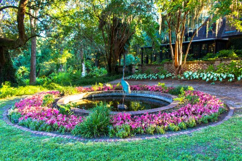 A circular garden pond surrounded by colorful flowers and lush greenery, with a sculpture in the center.