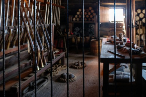 Interior of a rustic room with wooden shelves, tools, and barrels, viewed through metal bars.