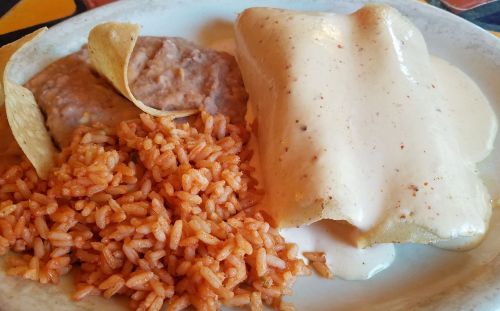 A plate of Mexican food featuring rice, refried beans, tortilla chips, and two enchiladas topped with creamy sauce.