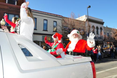 Santa Claus waves from the back of a truck during a festive parade, surrounded by holiday decorations and cheerful spectators.