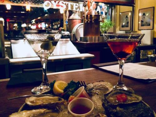 Two cocktails sit on a bar with a plate of oysters, lemon, and cocktail sauce in front of an oyster bar backdrop.