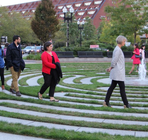 People walking through a grassy maze with a fountain in the background, surrounded by trees and buildings.