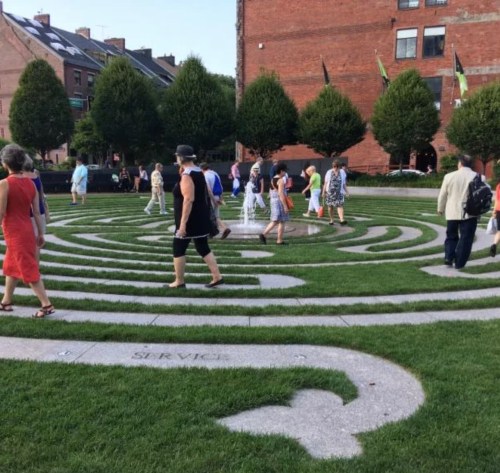 People walking through a grassy labyrinth in an urban setting, surrounded by trees and buildings.