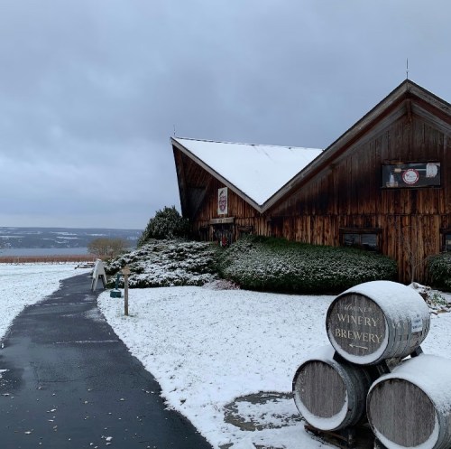 A rustic wooden building with a snowy landscape, featuring barrels in the foreground and a cloudy sky above.