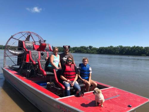 A group of four people and a dog on an airboat, enjoying a sunny day on the water.