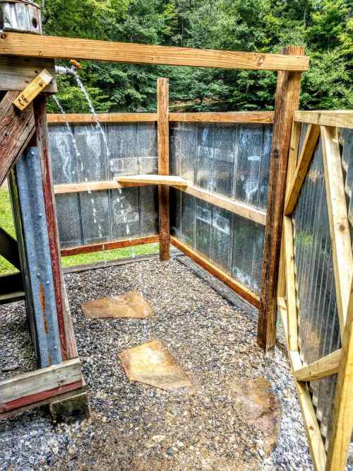 Outdoor shower area with wooden frame, gravel floor, and water flowing from a pipe, surrounded by metal panels.