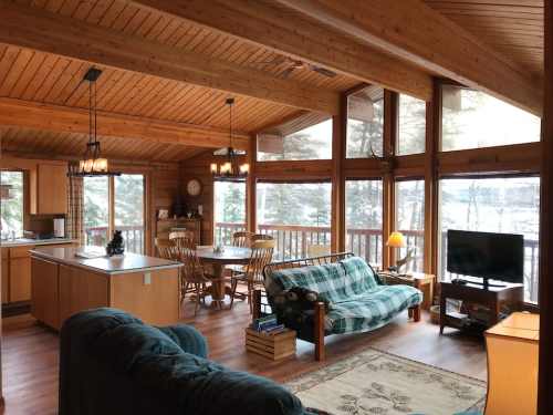 Cozy wooden cabin interior with a living area, dining table, and large windows overlooking a snowy landscape.