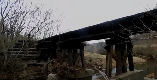 An old wooden bridge spans a dry riverbed, surrounded by bare trees and a hilly landscape.