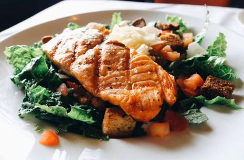 Grilled salmon fillet atop a fresh salad with greens, tomatoes, and croutons on a white plate.