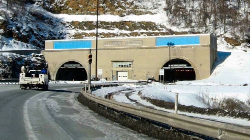 A snowy road leads to a large tunnel entrance with two arches, surrounded by rocky terrain and a clear blue sky.