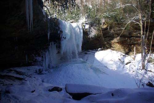 A frozen waterfall surrounded by snow-covered rocks and trees in a winter landscape.