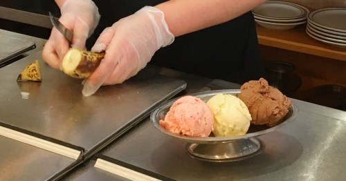 A person in gloves slices a banana next to a bowl of pink, yellow, and brown ice cream on a counter.