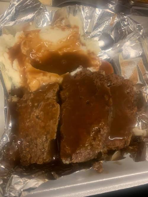 A serving of meatloaf with gravy, accompanied by mashed potatoes and brown gravy, in a foil container.