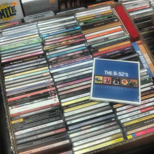A collection of CDs stacked in a box, featuring various album covers, with "The B-52's Original Album Series" prominently displayed.