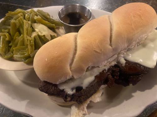 A sandwich with beef and melted cheese on a bun, served with green beans and a small cup of sauce on the side.