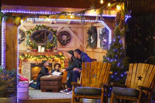 A family gathers in a cozy, decorated cabin, enjoying time together by a fireplace with festive lights.