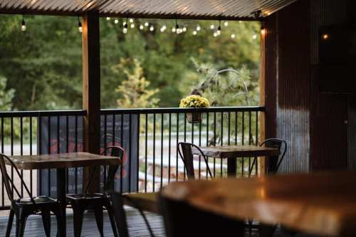 A cozy outdoor dining area with wooden tables, string lights, and a view of greenery in the background.