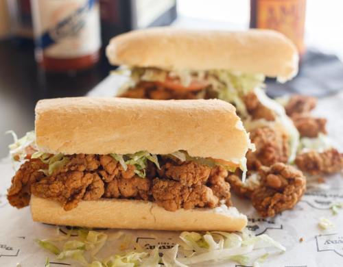 Two fried chicken sandwiches with lettuce and tomato, served on soft hoagie rolls, with drinks in the background.