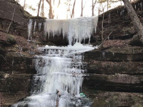 A frozen waterfall cascading down rocky cliffs, surrounded by trees and icicles, with sunlight peeking through.